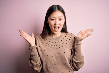 Genç, güzel Asyalı bir kadın pembe arka planda şık bir süveter giyerek çılgın ve başarıyı kollarını kaldırıp heyecanla haykırarak kutluyor. Kazanan konsept