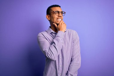 Yakışıklı Afrikalı Amerikalı adam mor arka plan üzerinde çizgili gömlek ve gözlük takıyor. Bir soru hakkında endişeleniyor, endişeli ve gergin.