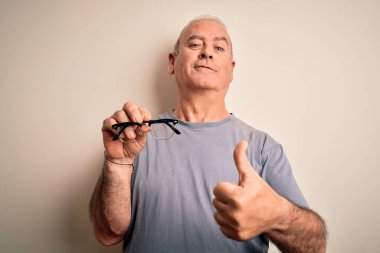 Orta yaşlı, beyaz arka planda görüş gözlüğü takan, beyaz gözlü, mutlu, gülümseyen, işaret veren, parmaklarıyla başparmağını kaldıran, mükemmel bir işaret.