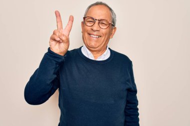 Üst düzey, gri saçlı, süveter ve gözlük takan, izole edilmiş beyaz arka planda gülümseyen, kameraya gülümseyen ve zafer işareti yapan bir adam. İki numara..