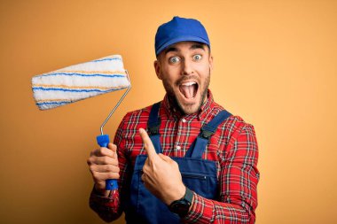 Genç yakışıklı ressam duvarları boyuyor. İzole edilmiş sarı arka plan üzerinde silindir kullanıyor. Elle ve parmakla göstererek çok mutlu.