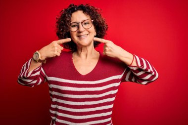 Orta yaşlı güzel kıvırcık saçlı kadın kırmızı arka planda sıradan çizgili süveter giyiyor neşeli gülüyor ve parmaklarıyla ve ağzıyla işaret ediyor. Diş sağlığı konsepti.