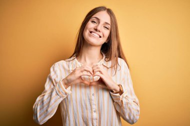 Genç, güzel, kızıl saçlı bir kadın, izole edilmiş sarı arka planda, gündelik çizgili tişört giyiyor. Sevgiyle gülümsüyor, elleriyle kalp sembolü yapıyor. Romantik konsept.
