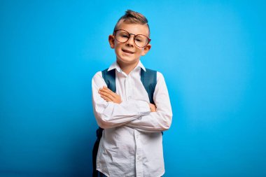 Zeki gözlüklü, mavi arka planda okul çantalı, kollarını kavuşturup kameraya gülümseyen küçük beyaz çocuk. Pozitif kişi.