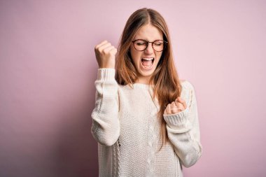 Genç, güzel, kızıl saçlı bir kadın pembe arka plan üzerine gündelik süveter ve gözlük takarak kolları açık ve gözleri kapalı bir şekilde başarıyı kutluyor. Kazanan konsept.