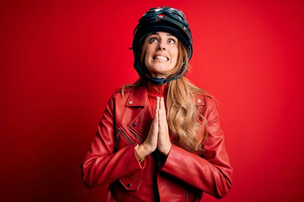 赤い背景を物乞いと手で祈る上で元ヘルメットを身に着けている若い美しいブルネットのオートバイの女性は 顔に希望の表現と一緒に非常に感情的かつ心配しています 物乞い — ストック写真