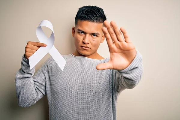 Young latin man holding white cancer ribbon supporting stop women violence with open hand doing stop sign with serious and confident expression, defense gesture