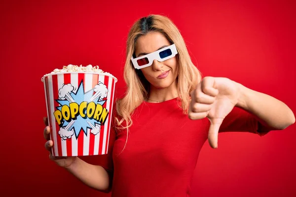 Young beautiful blonde woman watching movie using 3d glasses eating popcorns as snack with angry face, negative sign showing dislike with thumbs down, rejection concept