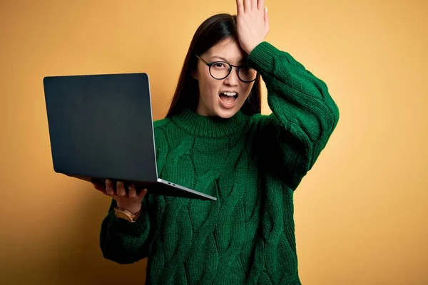 Young asian business woman wearing glasses and working using computer laptop surprised with hand on head for mistake, remember error. Forgot, bad memory concept.