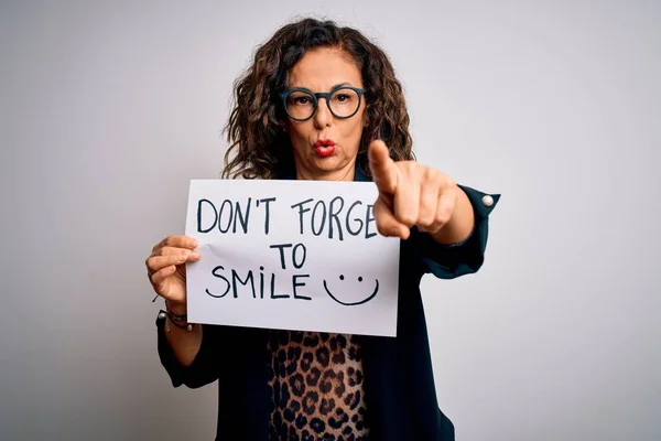 Middle age woman holding banner with dont forget smile message over white background pointing with finger to the camera and to you, hand sign, positive and confident gesture from the front