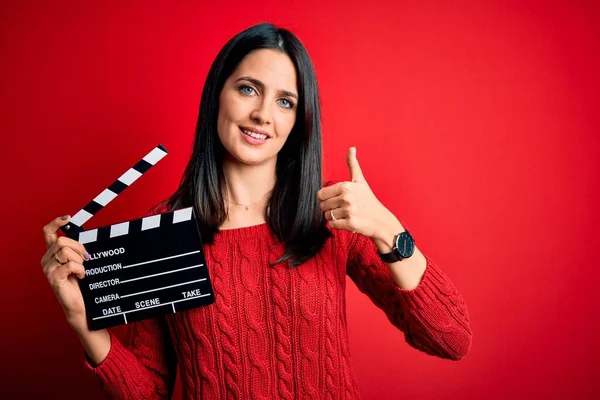 Young director woman with blue eyes making movie holding clapboard over red background happy with big smile doing ok sign, thumb up with fingers, excellent sign