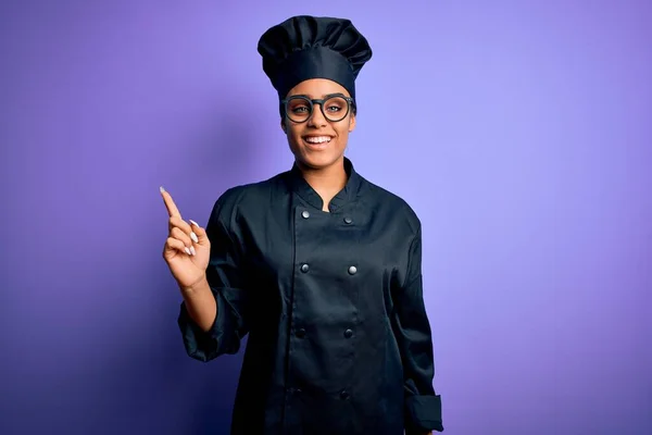 Young african american chef girl wearing cooker uniform and hat over purple background with a big smile on face, pointing with hand finger to the side looking at the camera.