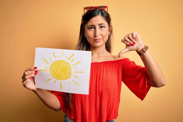 Young beautiful brunette woman holding paper with sun draw over isolated yellow background with angry face, negative sign showing dislike with thumbs down, rejection concept