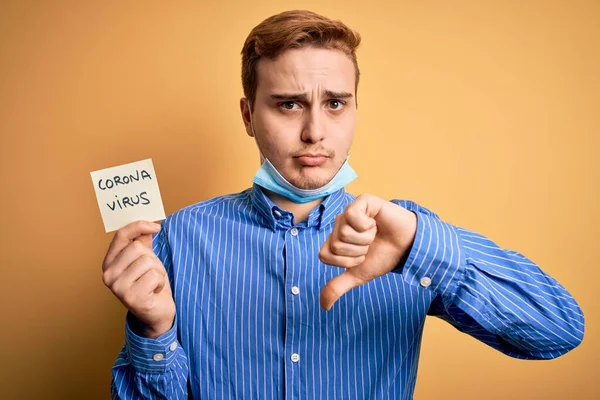 Young man wearing medical security mas holding paper note reminder with coronavirus alert word with angry face, negative sign showing dislike with thumbs down, rejection concept