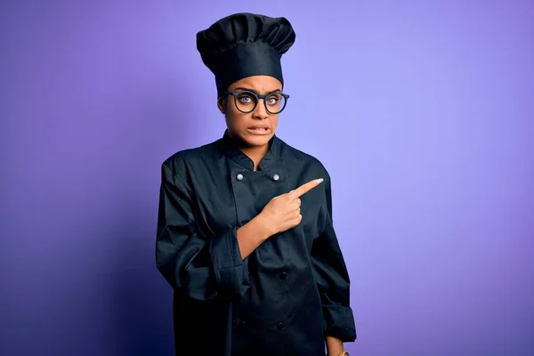 Young african american chef girl wearing cooker uniform and hat over purple background Pointing aside worried and nervous with forefinger, concerned and surprised expression