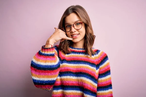 Young beautiful blonde girl wearing glasses and casual sweater over pink isolated background smiling doing phone gesture with hand and fingers like talking on the telephone. Communicating concepts.