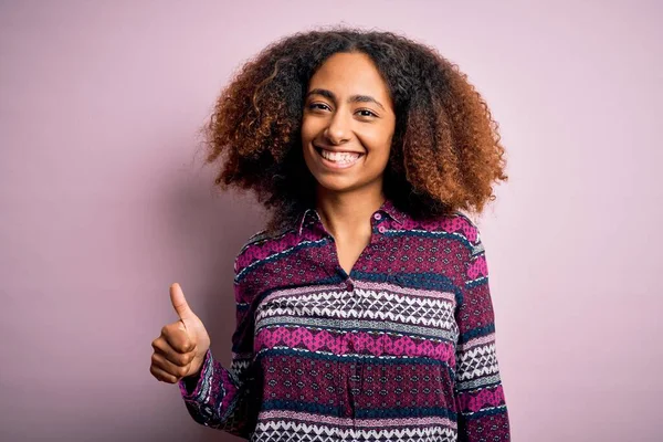 ピンクの背景にカラフルなシャツを着たアフロヘアの若いアフリカ系アメリカ人女性が手で幸せな親指アップジェスチャーをしています 成功を示すカメラを見て表現を承認する — ストック写真