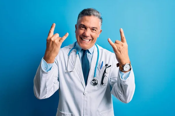 Middle age handsome grey-haired doctor man wearing coat and blue stethoscope shouting with crazy expression doing rock symbol with hands up. Music star. Heavy music concept.