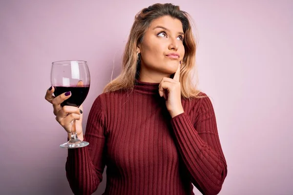若いです美しいですブロンド女性飲むガラスの赤ワイン以上隔離されたピンクの背景深刻な顔考えについて質問 非常に混乱したアイデア — ストック写真
