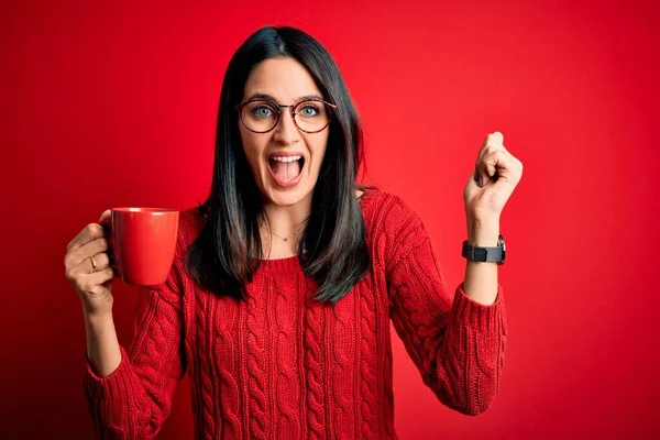 Young brunette woman with blue eyes wearing glasses and drinking a cup of coffee screaming proud and celebrating victory and success very excited, cheering emotion