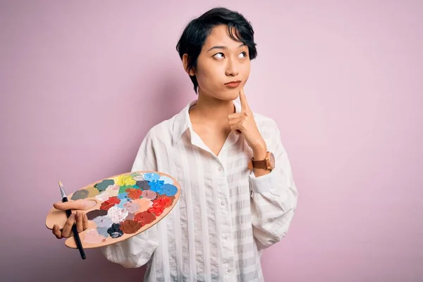 Young beautiful chinese artist woman painting using paintbrush and palette with colors serious face thinking about question, very confused idea