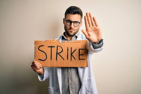 Young handsome doctor man protesting holding cardboard with strike message with open hand doing stop sign with serious and confident expression, defense gesture