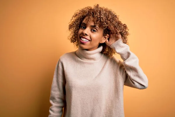 耳に噂やゴシップに耳を傾け手で笑みを浮かべて黄色の背景にタートルネックセーターを身に着けている若い美しいアフリカ系アメリカ人女性 病気の概念 — ストック写真