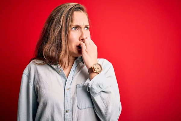 年轻漂亮的金发女人穿着休闲裤衬衫 披着孤立的红色背景 看上去很紧张 双手放在咬指甲的嘴上 焦虑问题 — 图库照片