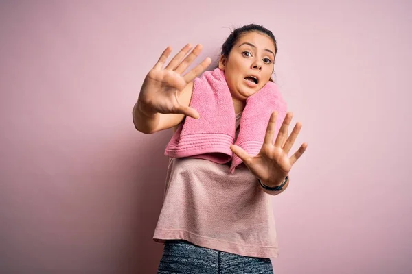 年轻美丽的黑发女运动员身穿运动服 披着粉色背景的毛巾 害怕恐惧地表情 用手停止手势 震惊地大叫 恐慌的概念 — 图库照片