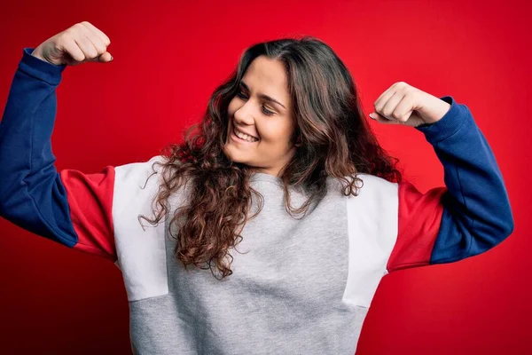 Young beautiful woman with curly hair wearing casual sweatshirt over isolated red background showing arms muscles smiling proud. Fitness concept.