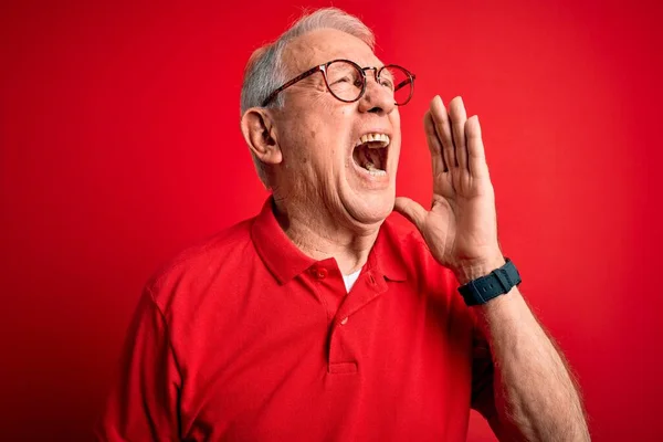 Grey haired senior man wearing glasses and casual t-shirt over red background shouting and screaming loud to side with hand on mouth. Communication concept.