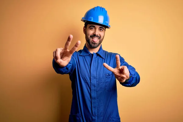機械的な男は 両方の手の勝利のサインを行う指を示す舌で笑顔黄色の背景に青い制服と安全ヘルメットを身に着けてひげを生やしている 第二番 — ストック写真
