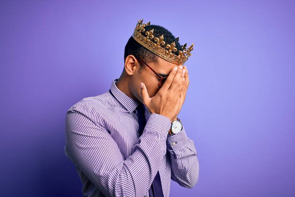 Young handsome african american man wearing golden crown of king over purple background with sad expression covering face with hands while crying. Depression concept.