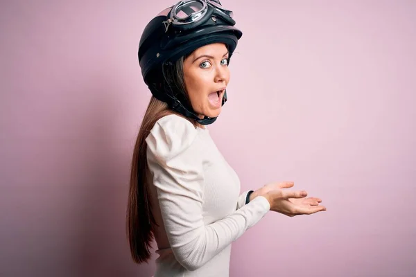 ピンクの背景に元ヘルメットを身に着けている青い目の若い美しいオートバイの女性は コピースペースを示す手を開いて手のひらで脇を指して 広告を提示笑顔興奮幸せな興奮 — ストック写真