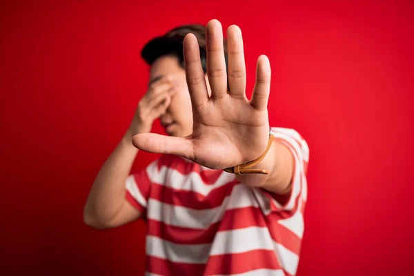 Young handsome chinese man wearing casual striped t-shirt standing over red background covering eyes with hands and doing stop gesture with sad and fear expression. Embarrassed and negative concept.