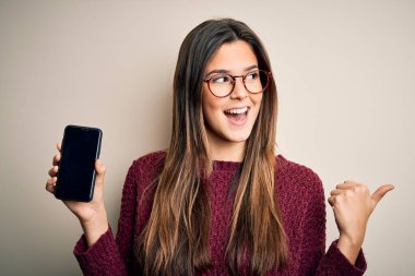 Gözlük takan genç güzel bir kız izole edilmiş beyaz arka plan üzerinde akıllı telefon gösteriyor ve gülümseyen bir gülümsemeyle baş parmağıyla işaret ediyor.