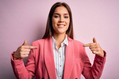 Güzel, esmer, ceketli bir kadın pembe arka planda duruyor. Yüzünde bir gülümsemeyle, gururlu ve mutlu bir şekilde kendini işaret ediyor..