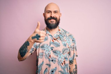 Sakallı ve dövmeli yakışıklı, pembe arka planda sıradan bir çiçek gömleği giyen, el hareketleriyle mutlu bir jest yapan kel bir adam. Kameraya bakıp başarıyı gösteren ifade onaylanıyor.