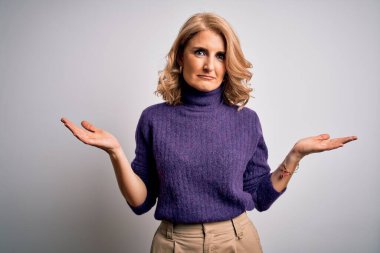 Beyaz arka plan üzerine mor balıkçı yaka kazak giyen orta yaşlı güzel bir kadın ve elleri ve kolları havada kafası karışmış bir ifade. Şüphe kavramı.