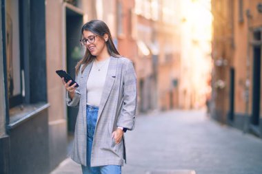 Mutlu ve kendinden emin gülümseyen genç ve güzel bir kadın. Yüzünde gülümsemeyle şehirde akıllı telefon kullanarak durmak.