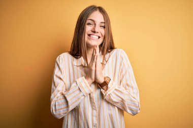 Genç, güzel, kızıl saçlı, günlük çizgili tişört giyen, izole edilmiş sarı arka planda dua eden ellerini birleştirip af dileyen, kendinden emin bir şekilde gülümseyen bir kadın..
