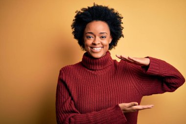 Kıvırcık saçlı, balıkçı yaka kazak giyen, elleri büyük ve büyük beden işareti gösteren genç, Afro-Amerikan bir kadın. Ölçü sembolü. Kameraya bakıp gülümsüyor. Ölçüm konsepti.