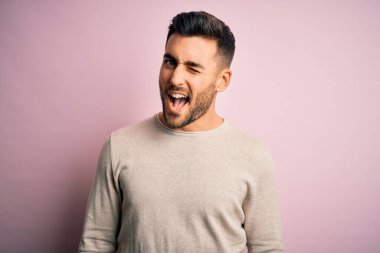 Genç yakışıklı bir adam, günlük süveter giyip pembe arka planda dikiliyor. Seksi bir ifadeyle kameraya bakıyor, neşeli ve mutlu bir yüzle..