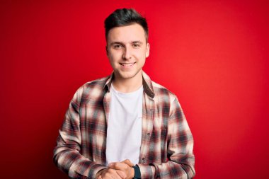 Kırmızı izole edilmiş arka plan üzerine gündelik modern tişört giyen genç, yakışıklı beyaz bir adam ellerini birleştirip gülümseyerek gevşemiş ve neşeli gülümsüyor. Başarı ve iyimser