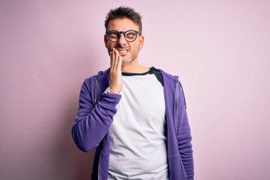 Mor eşofman altı giyen ve pembe arka planda duran gözlüklü genç bir adam diş ağrısı ya da diş ağrısı yüzünden acı dolu bir ifadeyle dudaklarına dokunuyor. Dişçi