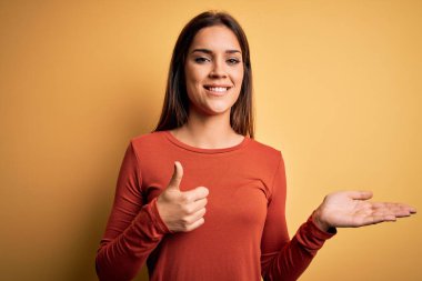 Genç, güzel, esmer, günlük tişört giyen, sarı arka planda duran, avuç içini gösteren, baş parmaklarıyla hareket eden, mutlu ve neşeli gülümseyen bir kadın.