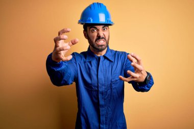 Sakallı tamirci, mavi üniformalı ve sarı arka planda kasklı. Öfkeden kudurmuş, elleri boğmaya çalışan, bağırıp çağıran biri.