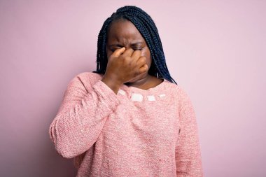 Siyah saçlı, uzun saçlı, pembe arka planda rahat süveter giyen, burnu ve gözleri ovuşturan yorgun ve başı ağrıyan Afro-Amerikan bir kadın. Stres ve hüsran kavramı.