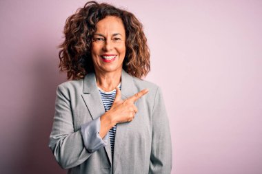 Orta yaşlı, güzel bir iş kadını zarif bir ceket giyiyor pembe arka planda neşeli bir yüzü var. Yüzünde mutlu ve doğal bir ifadeyle el ve parmak ile yüzünü gösteren bir gülümseme var.