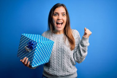 Sarışın genç bir kadın, izole edilmiş arka planda doğum günü hediyesi olarak bir hediye kutusu tutuyor. Gururla bağırıyor, zaferi ve başarıyı kutluyor. Çok heyecanlı, coşkulu bir duygu.